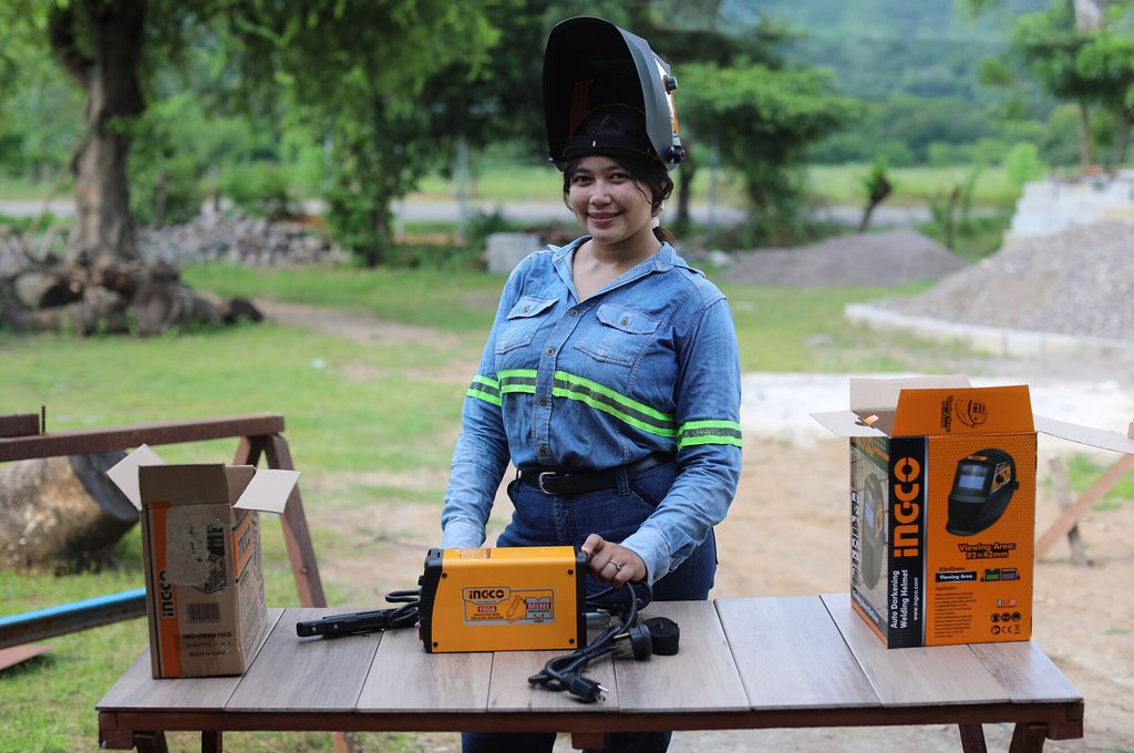 A young woman in protective gear stands at a table displaying assorted tools
